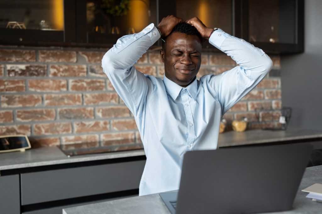 A frustrated young man sitting at a kitchen counter, holding his head in his hands with his eyes closed, looking stressed while using a laptop.