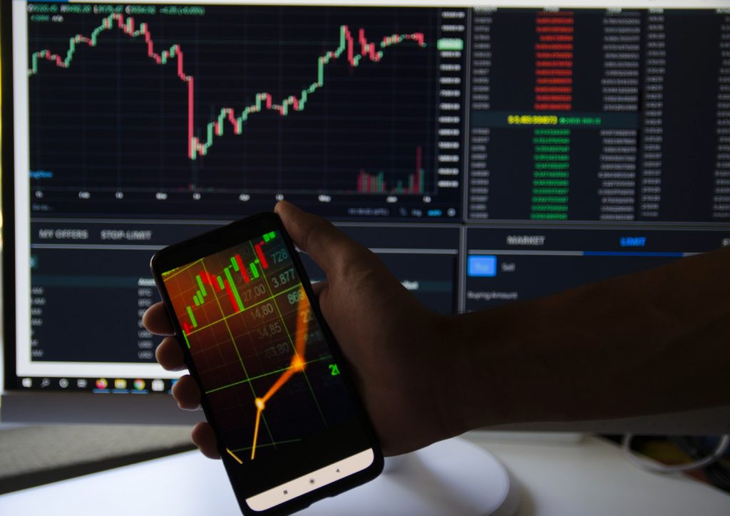 A person holding a smartphone displaying a crypto trading chart, with multiple trading screens in the background showing market trends and price movements.
