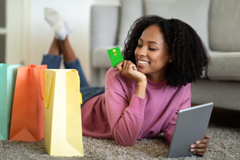 A smiling young woman lying on a carpet, holding a green credit card while shopping online with a tablet, surrounded by colorful shopping bags.