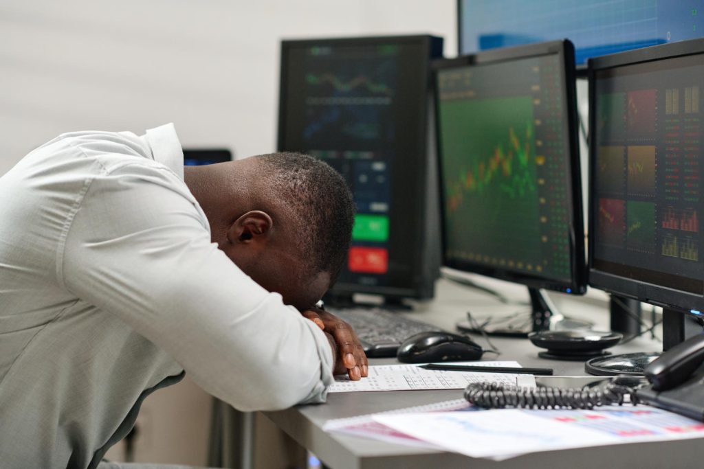 A frustrated crypto trader, sitting at their desk, leaning forward with their head down in disappointment. Their screens display a crypto chart with visible downward trends, symbolizing a loss in a trade. Pieces of paper are scattered around, adding to the sense of frustration